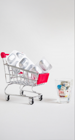 Descrição: Arte na qual vemos um carrinho de supermercado (em miniatura) com várias cartelas de comprimidos dentro. O fundo é liso e tem a cor branco gelo. 